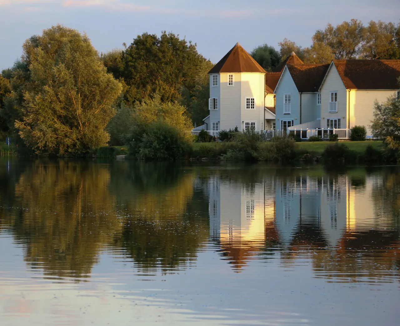 Grey Heron Lodge, a white Cape-Cod turret lodge, reflected in Windrush Lake at golden hour